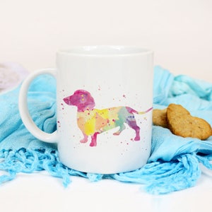 May include: White ceramic mug featuring a watercolour dachshund silhouette in shades of pink, yellow, and green. The dachshund is facing left and has a long tail.