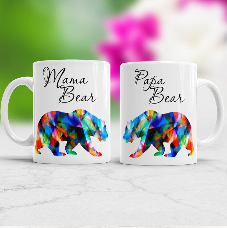 May include: Two white ceramic mugs with black text and colorful bear silhouettes. The mug on the left says "Mama Bear" and the mug on the right says "Papa Bear".