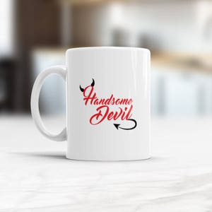 May include: White ceramic coffee mug with a black devil tail and horns design. The text "Handsome Devil" is written in red cursive.