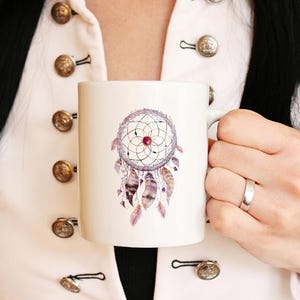 May include: White ceramic mug with a watercolor design of a dreamcatcher with purple, pink, and brown feathers. The dreamcatcher has a red bead in the center.