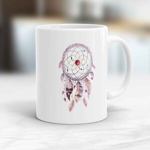 May include: White ceramic mug with a watercolor illustration of a dreamcatcher with feathers and a red gemstone in the center.