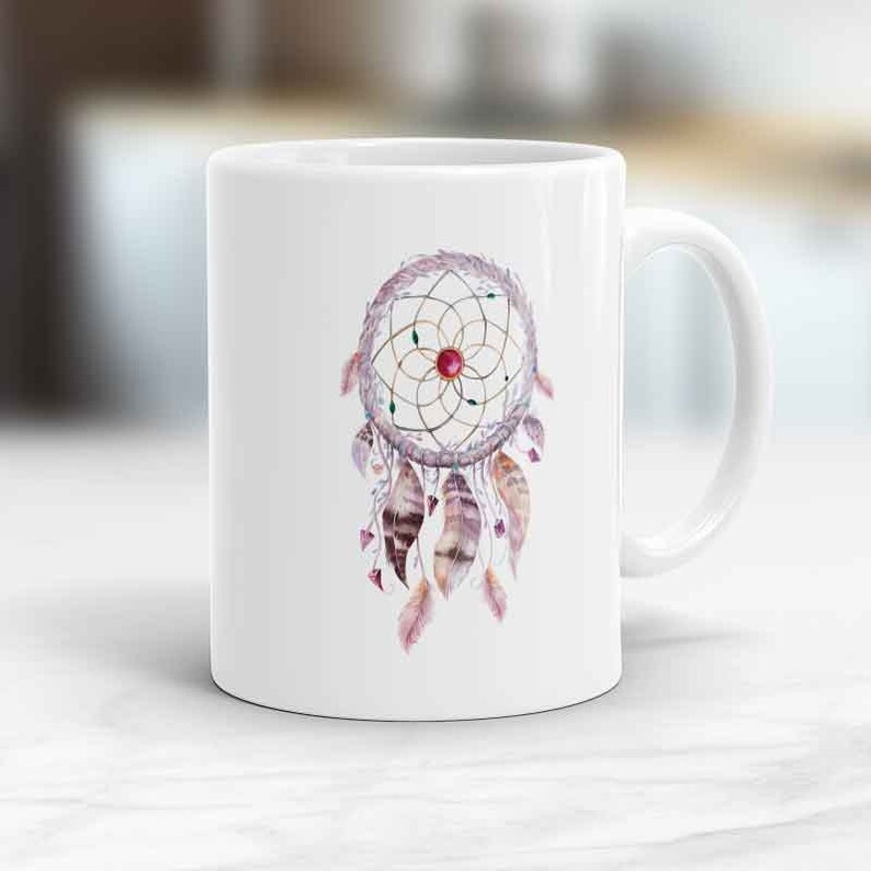 May include: White ceramic mug with a watercolor illustration of a dreamcatcher with feathers and a red gemstone in the center.