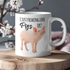 Könnte beinhalten: Weiße Keramiktasse mit Henkel. Auf der Tasse steht der Text "I JUST FREAKING LOVE Pigs OK?" in schwarzer Schrift und eine rosa Schweineillustration. Die Tasse steht auf einer Holzoberfläche.