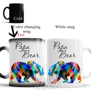 May include: A color-changing mug with a black exterior and a white interior. The mug features the text "Papa Bear" in black cursive script and a colorful bear design. The mug changes color when hot liquid is poured into it.