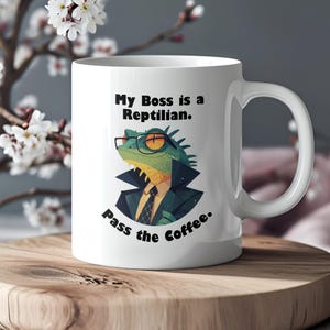 May include: White ceramic mug with a black and green illustration of a lizard wearing glasses and a suit. The text on the mug reads "My Boss is a Reptilian. Pass the Coffee."