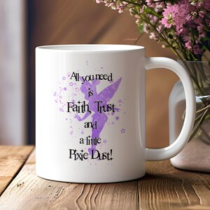 May include: White ceramic mug with a purple Tinkerbell silhouette and the text "All you need is Faith, Trust and a little Pixie Dust!" The mug has a curved handle and is set on a wooden surface.