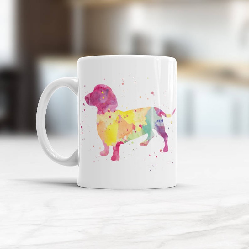 May include: A white ceramic mug featuring a colourful watercolour silhouette of a dachshund. The dachshund is pink, yellow, orange, and blue. The mug has a white handle.
