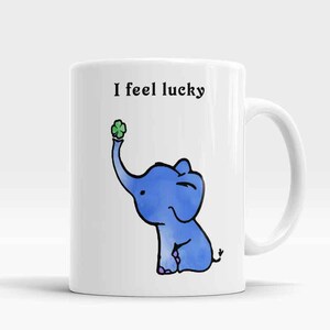 Op de afbeelding: Een witte keramische mok met een blauwe olifant die een klavertje vier vasthoudt. Op de mok staat de tekst "I feel lucky".