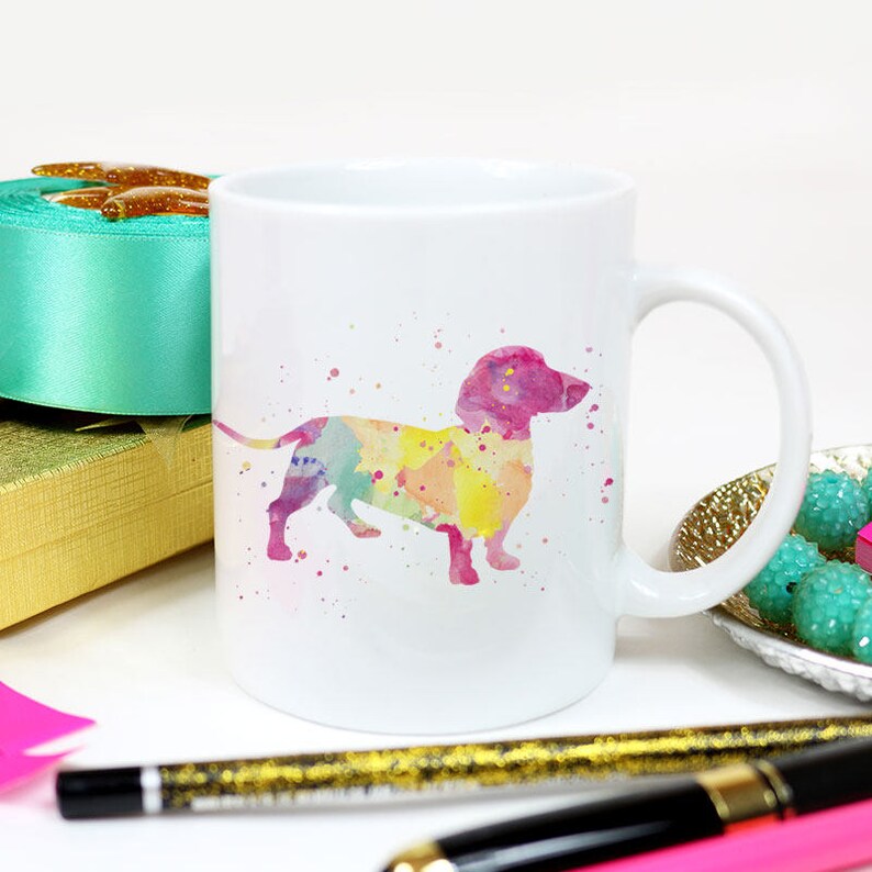 May include: White ceramic coffee mug featuring a watercolour dachshund silhouette in shades of pink, yellow and blue.