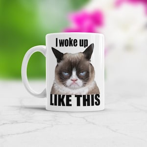 Pode incluir: Caneca de café de cerâmica branca com uma imagem de um gato mal-humorado e o texto "I woke up like this".