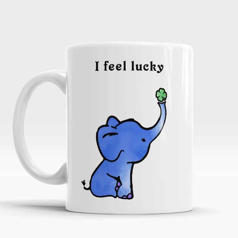 Op de afbeelding: Een witte keramische mok met een illustratie van een blauwe olifant die een groen klavertje vier houdt. Op de mok staat de tekst "I feel lucky".