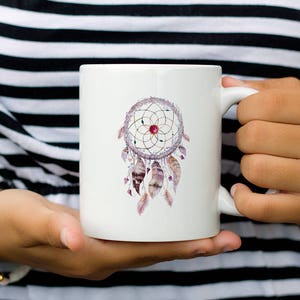 May include: White ceramic mug with a watercolor dreamcatcher design. The dreamcatcher is pink, purple, and gold with feathers.