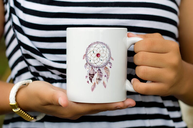 May include: White ceramic mug with a watercolor dreamcatcher design. The dreamcatcher is pink, purple, and gold with feathers.