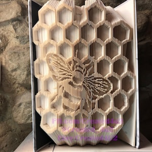 May include: A book folded into a three-dimensional honeycomb design with a bee in the center. The book is standing upright on a stack of books.