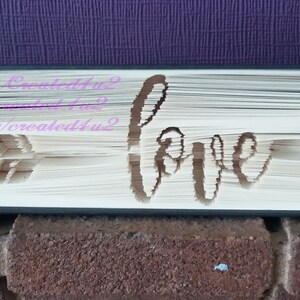 May include: A book with the word "love" cut out of the pages. The book is standing on a brick surface. The book is decorated with an arrow on the left side and the text "FB Created4u2 Etsy/created4u2" on the right side.