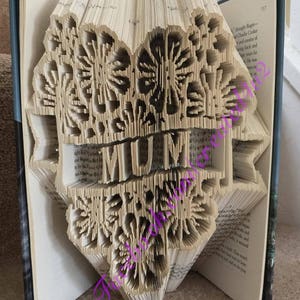 Könnte beinhalten: Eine Buchkunstskulptur des Wortes "MUM", die aus den Seiten eines Buches gefertigt ist. Die Skulptur besteht aus gefalteten Seiten und hat ein florales Design.