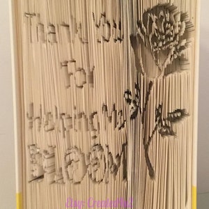 Peut inclure: Un livre dont les pages sont pliées pour créer un design 3D d'une rose et les mots "Merci Maman". Le livre a des bords jaunes et le texte "Etsy: Created4U FB: Created4U @Created4U" en bas.