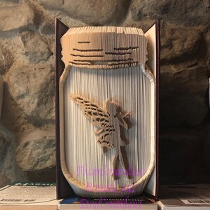 May include: A book with a fairy cut out of the pages, shaped like a mason jar. The book is standing upright on a stack of books. The book is brown with a purple spine.
