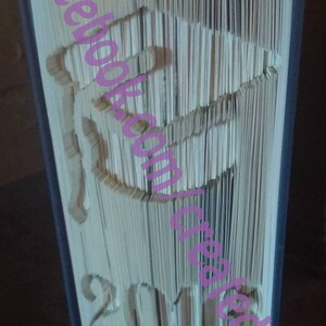 May include: A handcrafted book with pages intricately cut to reveal a graduation cap design and the year 2018. The book's edges are painted blue, and the text "facebook.com/create4u2" is overlaid in pink.
