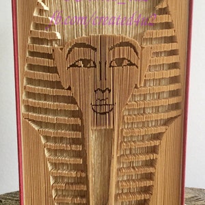 May include: A book art sculpture of an Egyptian pharaoh's head, created by folding the pages of a book. The sculpture is made of brown paper and is displayed on a table with a patterned surface.