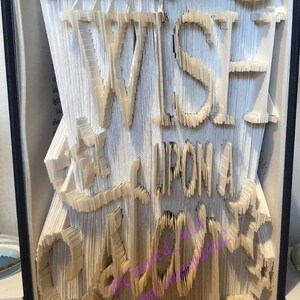 May include: A book with pages intricately folded and cut to reveal the words "WHEN YOU WISH UPON A STAR". The text is in a whimsical, stylized font, creating a unique piece of book art. The book's edges are visible.