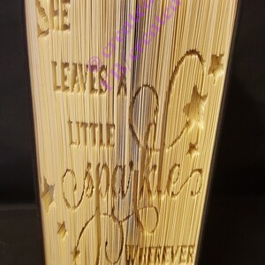 May include: A book with pages carved to read "She leaves a little sparkle wherever she goes." The text is cut into the pages, with star and heart accents. The book has a black spine and is set against a dark background.