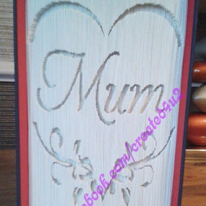 May include: A white book with a heart shape cut out and the word "Mum" inside. The heart is decorated with a floral design. The book has red edges.