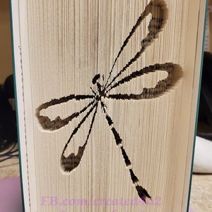 May include: A book art sculpture of a dragonfly, created by folding the pages of a book. The dragonfly is cut out of the pages, creating a 3D effect. The book is bound in a green cover.