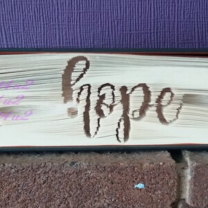 May include: A book with the word "hope" cut out of the pages. The pages are folded to create a three-dimensional effect. The book is sitting on a brick surface.