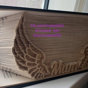 May include: A book with pages folded into a 3D design of wings and the word "LOVE". The book is closed and the spine is visible. The book is on a white surface.