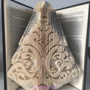 Può includere: Una scultura di libro con intricati disegni ritagliati, che formano una forma tridimensionale. Le pagine sono beige e il libro è aperto, rivelando i dettagli scolpiti. La scultura ha un tema decorativo e ornamentale, con testo visibile sulle pagine. Gli URL di Etsy e Facebook sono visibili.