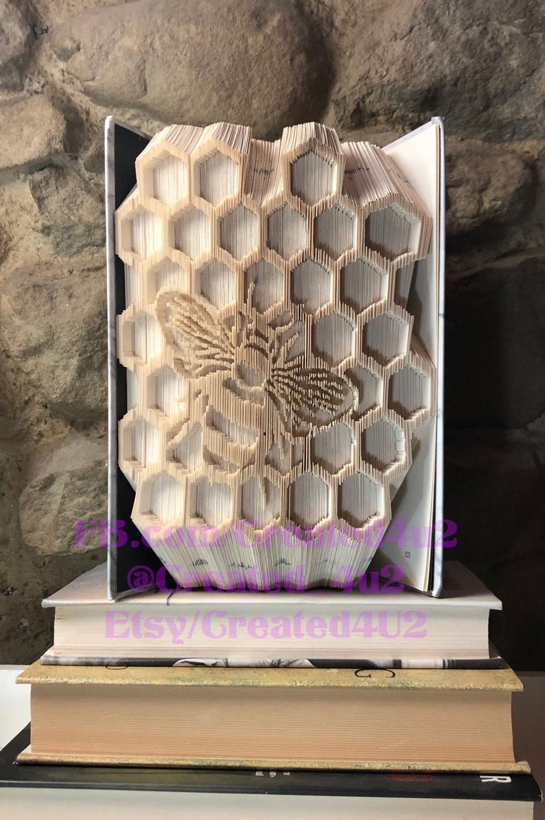 May include: A book art sculpture of a honeycomb with a bee in the center. The book is folded into a three-dimensional shape and is displayed on a stack of books. The book art is made from a book with a white cover.