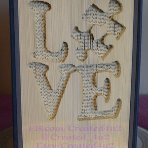 May include: A book art sculpture of the word "LOVE" created by folding the pages of a book. The sculpture is displayed on a round, silver-colored base. The text "FB.com/Created4u2 @Created_4u2 Etsy/Created4u2" is printed on the bottom of the sculpture.