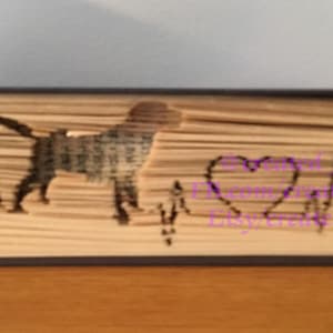 Puede incluir: Un libro con una silueta de perro y un corazón con una línea de latido del corazón tallada en las páginas. El texto "Forever in my heart" también está tallado en las páginas.
