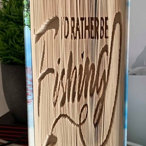 May include: A book with the words "I'd rather be fishing" carved into the pages. The book is standing upright on a table. The cover of the book is blue and white with a picture of a lake.