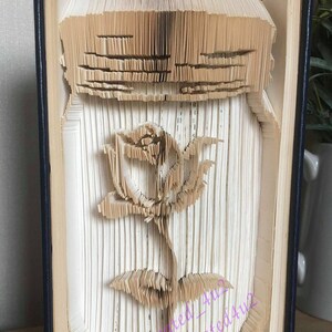 May include: A book with pages folded into the shape of a glass jar with a rose inside. The book is bound in blue and the rose is made of paper.