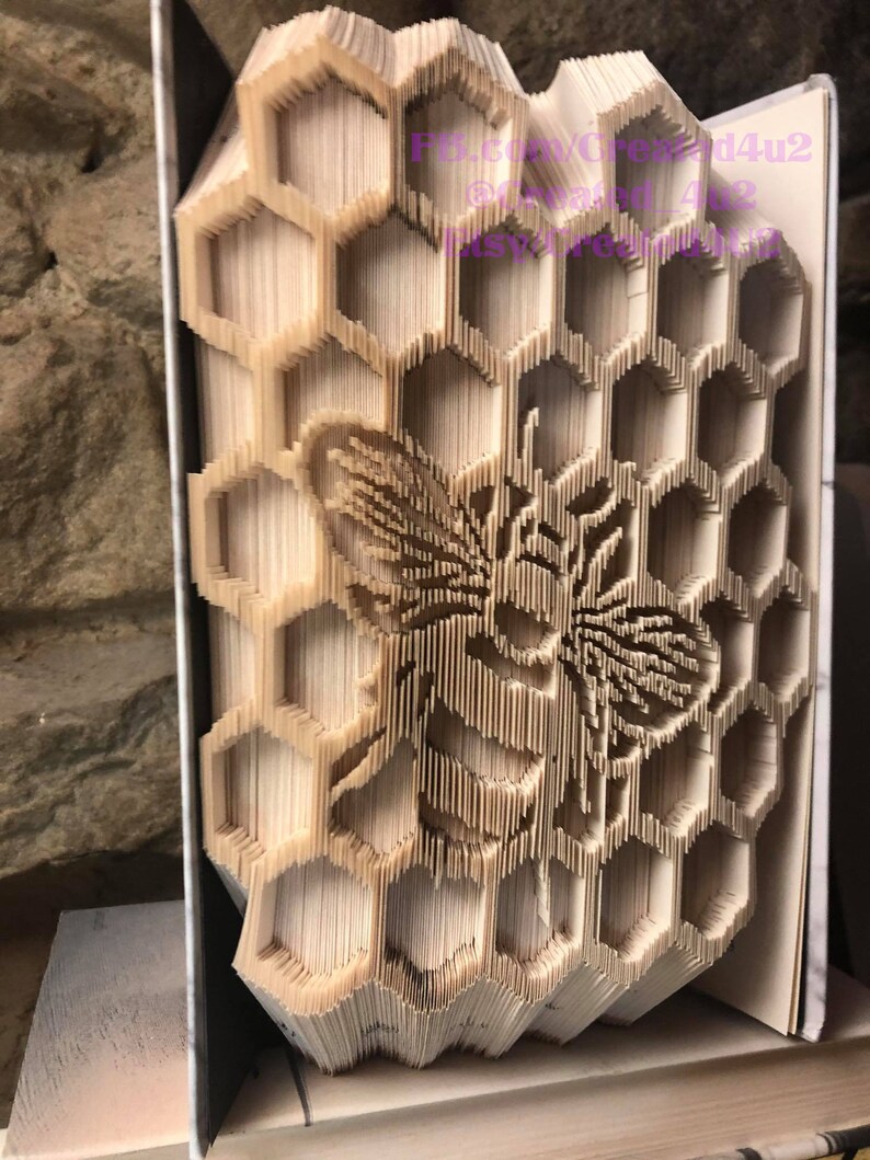 May include: A book folded into a 3D sculpture of a honeycomb with a bee in the center. The sculpture is made of paper and is a light brown color.