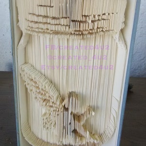 Può includere: Una scultura di arte libraria di un barattolo di muratore con una libellula all'interno. Il libro è piegato e modellato per creare un effetto 3D. La scultura è realizzata dalle pagine di un libro.