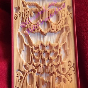 May include: A book with pages folded into the shape of an owl. The owl is made of beige paper and has a detailed design. The book is closed and is sitting on a red surface.