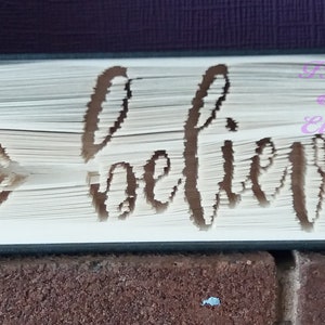 May include: A book with the word "believe" cut out of the pages, with an arrow on the left side. The book is bound in black and has a white spine.