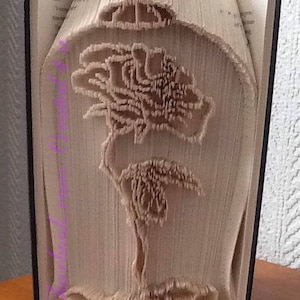 May include: A book folded into the shape of a rose, with a glass dome on top. The rose is made of paper and is a light brown color.