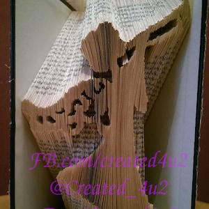May include: A book art sculpture of a tree, created by folding the pages of a book. The book is bound in black and the pages are folded to create a three-dimensional tree shape. The sculpture is displayed on a wooden surface. The text "FB.com/created4u2 @created_4u2 Etsy/created4u2" is visible on the book.