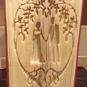 Op de afbeelding: Een boek met pagina's die zijn gevouwen tot een driedimensionaal ontwerp van een stel dat onder een boom staat. Het boek is gebonden in rood en de pagina's zijn lichtbruin.