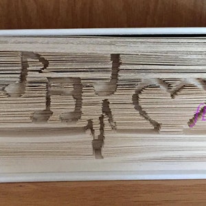 Puede incluir: Un libro con una cubierta blanca se ha doblado para crear un diseño tridimensional de una nota musical, un corazón y la palabra "Música".