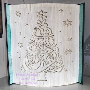 May include: A white book with a 3D pop-up Christmas tree design. The tree is decorated with snowflakes and swirls. The book is closed and standing upright on a table.