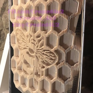 May include: A book folded into a 3D honeycomb design with a bee in the center. The book is standing upright on a table.