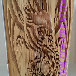 May include: A book sculpture of a dragon, created by folding the pages of a book. The sculpture is made from a brown book with a white spine. The dragon is facing the right side of the image. The book is standing on a white surface. The text "FACEBOOK.COM/BOOKCREATIONS" is visible on the right side of the book.