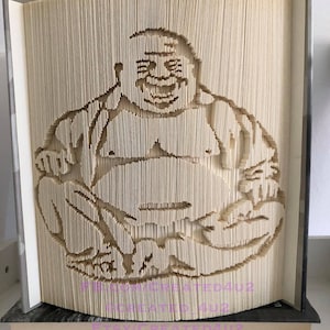 May include: A beige book with a 3D paper sculpture of a smiling, round-faced Buddha sitting cross-legged. The sculpture is made of folded paper and is a light brown color.