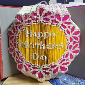 Może przedstawiać: Żółta książka ze słowami "Happy Mothers Day" złożona w trójwymiarowy wzór. Wzór otoczony jest różowo-białym kwiatowym obramowaniem.