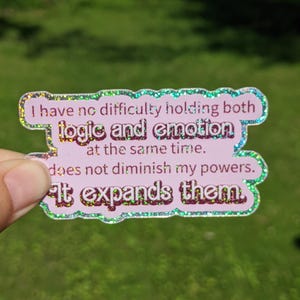 May include: A pink sticker with glitter edges and text that reads, "I have no difficulty holding both logic and emotion at the same time. It expands them." The sticker is held against a green background.